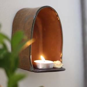May include: A brown ceramic wall sconce with a teal rim, holding a lit tea light candle and a small seashell. The interior of the sconce glows with the candle's light, creating a warm ambiance. A decorative item.