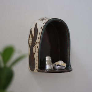 May include: A small, ceramic wall shelf with a dark brown interior and a decorative exterior. The shelf is adorned with brown leaf-like patterns and holds two rings and a small silver thimble. The shelf is mounted on a white wall.