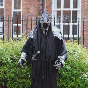 Può includere: Una persona in costume nero con elmo e spalline argentate, che tiene una spada. Il costume ha una lunga veste fluente e guanti. Lo sfondo include un edificio in mattoni e una recinzione nera.