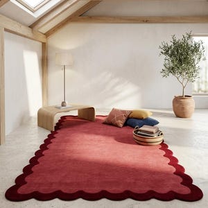 May include: A large, scalloped-edge rug in a deep red hue dominates the room. The rug has a darker red border. A wooden coffee table, pillows, and a woven basket with books are on the rug. A potted olive tree and a floor lamp are also in the room.