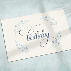 May include: A cream-colored birthday card with the words "H.A.P.P.Y Birthday" in elegant dark blue script. The card is adorned with delicate blue floral accents, creating a soft, celebratory design. The card is on a light blue surface.