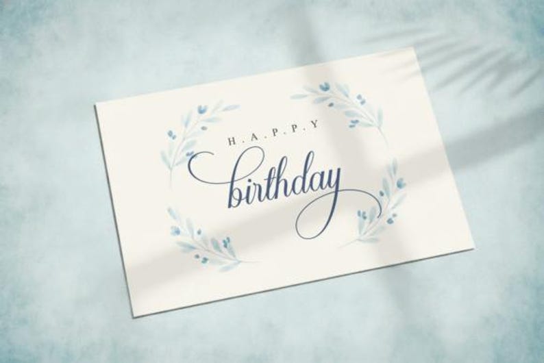May include: A cream-colored birthday card with the words "H.A.P.P.Y Birthday" in elegant dark blue script. The card is adorned with delicate blue floral accents, creating a soft, celebratory design. The card is on a light blue surface.