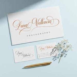 May include: A stationery set featuring a business card, two smaller cards, and a larger card with the name "Luna Valencia Photography" in elegant gold script. A gold pen and sprig of white flowers are also present.