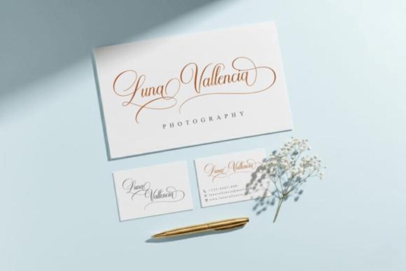 May include: A stationery set featuring a business card, two smaller cards, and a larger card with the name "Luna Valencia Photography" in elegant gold script. A gold pen and sprig of white flowers are also present.