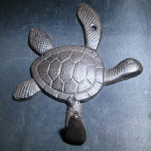 May include: A cast iron turtle-shaped wall hook in a dark bronze finish. The hook features a detailed turtle design with a textured shell and flippers. The hook is designed to be mounted on a wall for hanging items.