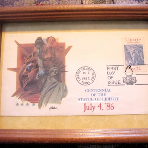 May include: A framed first day of issue stamp commemorating the centenary of the Statue of Liberty. The stamp features a blue and white illustration of the Statue of Liberty. The text "Liberty Island, NY, Jul 4, 1986, 10004" is printed on the stamp. The text "First Day of Issue" is printed below the stamp. The text "Centenary of the Statue of Liberty" is printed below the stamp. The text "July 4, '86" is printed below the stamp.