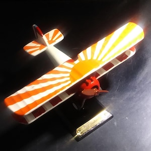 May include: A red and white model airplane with a rising sun design on the wings. The airplane is on a black stand.