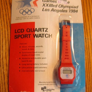 May include: A red and white LCD quartz sports watch with a red strap. The watch face displays the time and date. The packaging features the 1984 Los Angeles Olympics logo and the text "Games XXIIIrd Olympiad Los Angeles 1984".