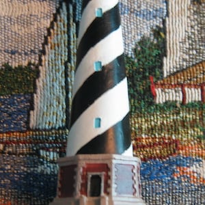 Puede incluir: Una figurita de faro de rayas blancas y negras con un techo azul y una base verde que dice "Cape Hatteras".