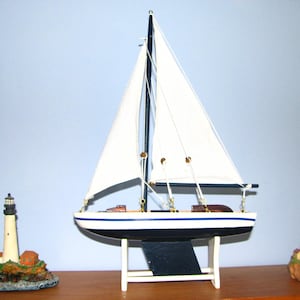 May include: A model sailboat with a dark blue hull, white upper section, and white fabric sails, supported by a white stand. Flanking the sailboat are two miniature lighthouse figurines, one on the left and one on the right, each featuring a tower.