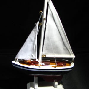 May include: A model sailboat featuring a dark blue hull with a white stripe, a wooden deck, and two white fabric sails attached to a central mast. The vessel is mounted on a white stand and includes a red keel visible beneath the hull structure.
