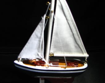 May include: A model sailboat featuring a dark blue hull with a white stripe, a wooden deck, and two white fabric sails attached to a central mast. The vessel is mounted on a white stand and includes a red keel visible beneath the hull structure.