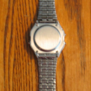 Puede incluir: Un reloj plateado con una esfera rectangular y una correa de metal a juego. El reloj se muestra sobre un fondo de madera. La correa est&aacute; hecha de peque&ntilde;os eslabones rectangulares conectados.