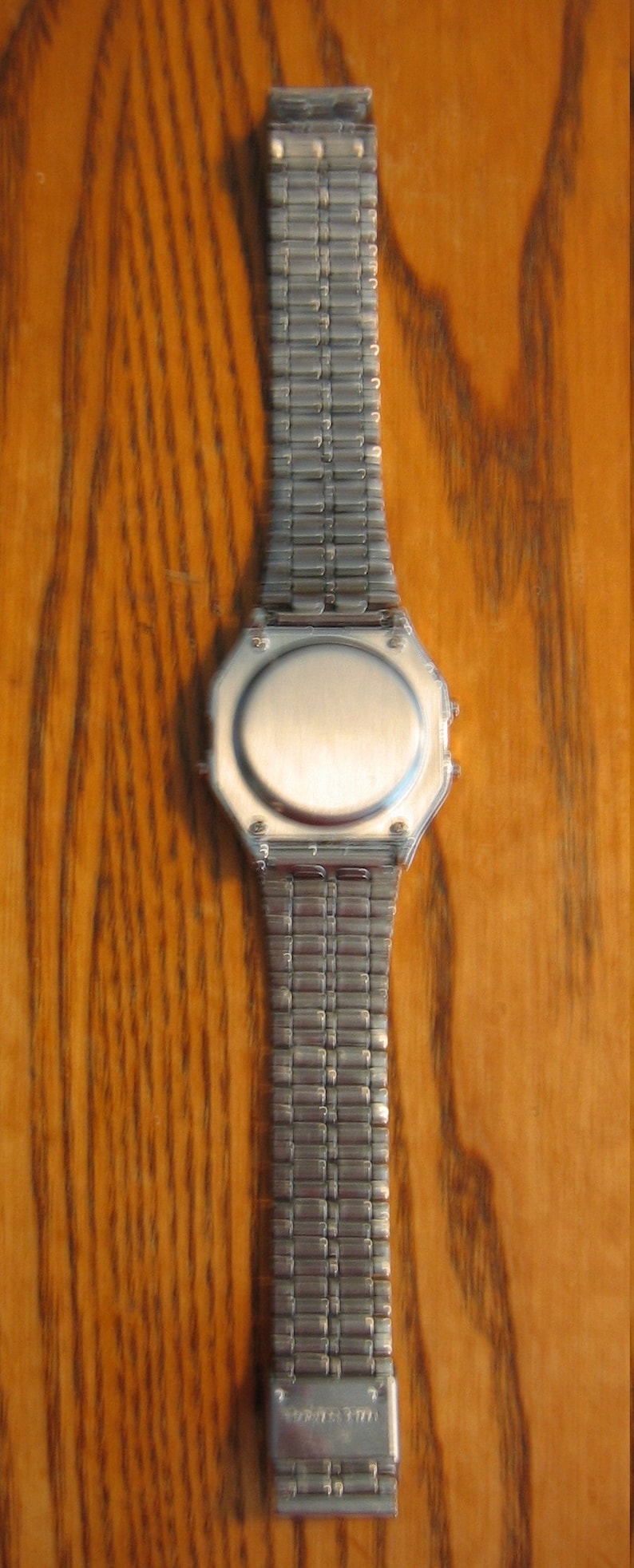 Puede incluir: Un reloj plateado con una esfera rectangular y una correa de metal a juego. El reloj se muestra sobre un fondo de madera. La correa est&aacute; hecha de peque&ntilde;os eslabones rectangulares conectados.