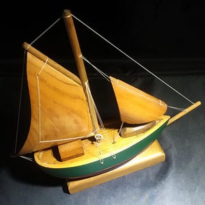 May include: A wooden model sailboat with two sails, a green hull, and a wooden base. The sails are made of wood and are painted a light brown colour. The hull is painted a dark green colour with a red stripe around the middle. The base is made of wood and is painted a light brown colour.