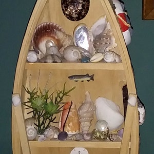 Puede incluir: Una estantería de madera con forma de barco con tres estantes, decorada con conchas marinas, un pequeño faro, un pez y un pequeño velero. Los estantes están llenos de varias conchas marinas y otras decoraciones náuticas.