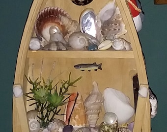 May include: A wooden boat-shaped shelf with three shelves, decorated with seashells, a small lighthouse, a fish, and a small sailboat. The shelves are filled with various seashells and other nautical decorations.