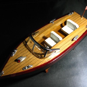 以下が含まれることがあります： 赤と茶色の船体、白い内装、後ろに小さなアメリカ国旗が付いた木製模型のボート。ボートにはフロントガラスと3つの座席があります。