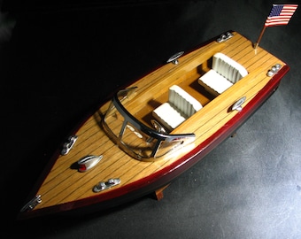 May include: A wooden model boat with a red and brown hull, a white interior, and a small American flag on the back. The boat has a windscreen and three seats.
