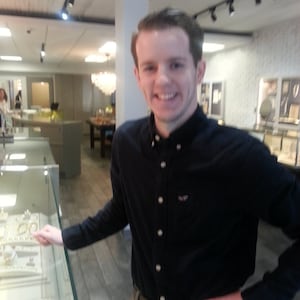 Puede incluir: Un hombre con una camisa negra y pantalones color caqui est&aacute; en una joyer&iacute;a. La tienda tiene vitrinas de cristal llenas de joyas. El hombre sonr&iacute;e y se&ntilde;ala las joyas.