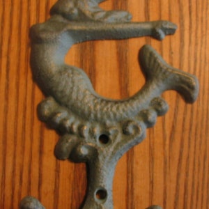 May include: A teal blue cast iron mermaid hook with two hooks at the bottom. The mermaid is depicted with a flowing tail and a whimsical pose.