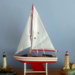 May include: A red and white wooden sailboat model with white sails and a red mast. The model is displayed on a white stand. Two miniature lighthouse figurines are on either side of the sailboat.