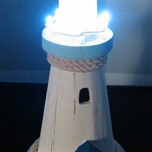 Może przedstawiać: Dekoracyjna lampa w kształcie latarni morskiej z niebieskim dachem i podstawą, białym korpusem i akcentem z liny. Lampa ma dołączony mały domek i emituje jasne białe światło. Latarnia morska to element wystroju domu o tematyce żeglarskiej.