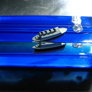 Könnte beinhalten: Ein blauer Glasblock mit zwei kleinen schwarzen Booten darin. Die Boote schwimmen auf dem Wasser, das klar und blau ist. Der Block steht auf einer schwarzen Oberfläche.
