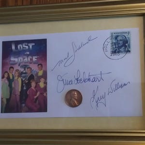May include: A framed photo of the cast of the television show "Lost in Space" with autographs from the actors. The signatures include: "Mark Goddard", "June Lockhart", and "Guy Williams". A 1968 US postage stamp with a portrait of George Washington is in the upper right corner.
