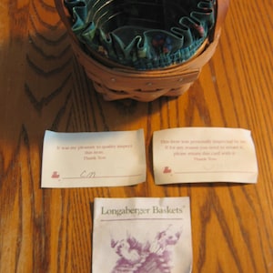 May include: A Longaberger basket with a green and floral fabric liner. The basket is made of woven wood and has a handle. The image also includes two cards with text that reads "It was my pleasure to quality inspect this item. Thank You" and "This item was personally inspected by me. If for any reason you need to return it, please return this card with it. Thank You".