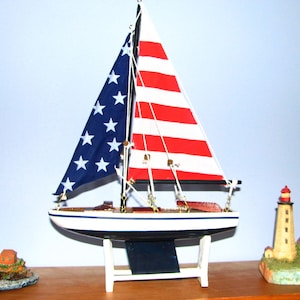 May include: A decorative sailboat model with a blue sail featuring white stars and a red and white striped sail. The boat is white with a black keel and sits on a white stand. Two lighthouse figurines flank the boat, creating a nautical theme.
