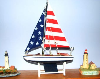 May include: A model sailboat with a blue sail with white stars and a red and white striped sail. The boat is white with a black keel and sits on a white stand. Two lighthouse ornaments flank the boat, creating a nautical theme.