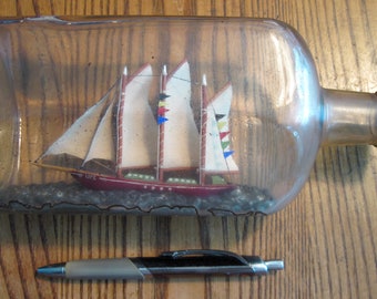 Op de afbeelding: Een miniatuur houten zeilboot met witte zeilen en rode, blauwe en gele vlaggen bevindt zich in een heldere glazen fles met een zwarte plastic dop. De fles staat op een houten oppervlak.