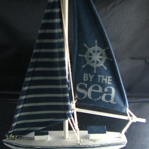 May include: A miniature sailboat model with a white hull and blue sails. One sail has horizontal white stripes, the other has a ship's wheel and the text "BY THE sea". The boat is mounted on a white stand.