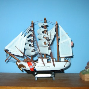 May include: A model ship of the US Coast Guard, with white sails and a red, white, and blue flag. The ship is on a wooden shelf with a lighthouse model on either side.