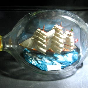May include: A glass bottle with a miniature sailing ship inside. The ship features white sails and is set on a blue base, simulating water. The bottle's neck is sealed with a rope-bound cork.