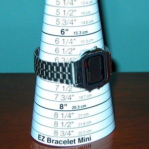 Puede incluir: Un reloj digital plateado se muestra en un cono de medici&oacute;n blanco EZ Bracelet Mini. El cono tiene medidas en pulgadas y cent&iacute;metros, que van desde 5 pulgadas (12,7 cm) hasta 8 3/4 pulgadas (22,2 cm).