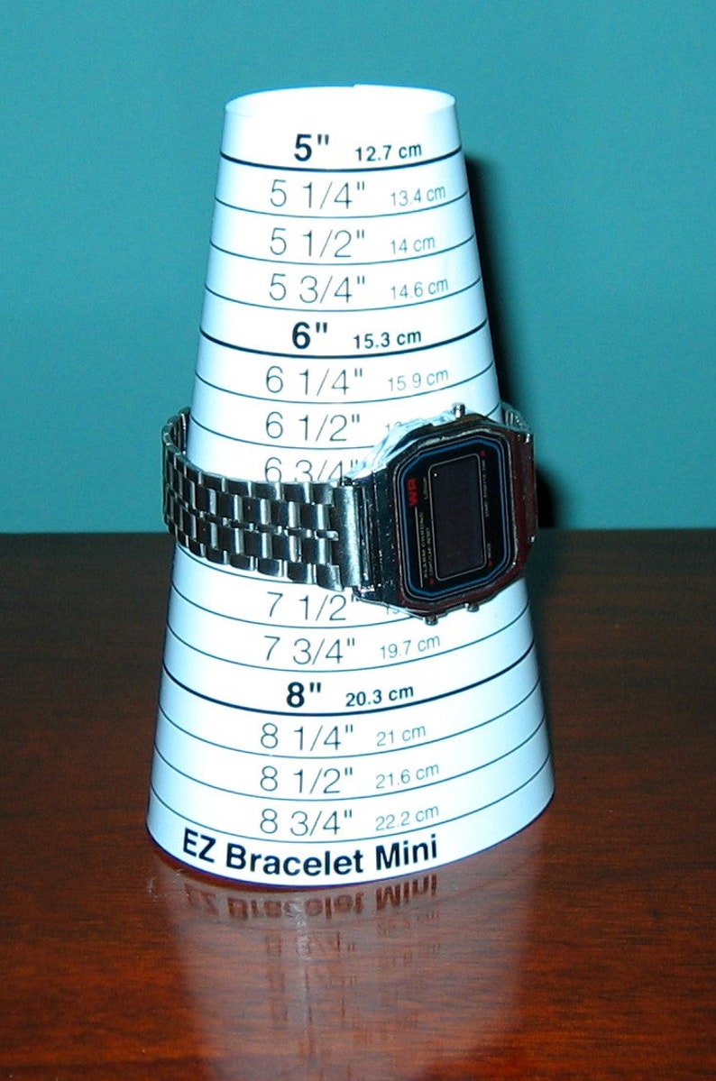 Puede incluir: Un reloj digital plateado se muestra en un cono de medici&oacute;n blanco EZ Bracelet Mini. El cono tiene medidas en pulgadas y cent&iacute;metros, que van desde 5 pulgadas (12,7 cm) hasta 8 3/4 pulgadas (22,2 cm).