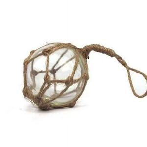 Puede incluir: Un flotador de pesca de vidrio transparente, envuelto en una red de cuerda marrón tejida. El flotador tiene un lazo de cuerda adjunto para colgar. El artículo decorativo de temática náutica está sobre un fondo blanco.