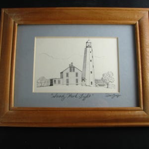 May include: Framed black and white drawing of a lighthouse and adjacent building, set against a light blue background. The artwork is in a wooden frame. The text "Sand Hook Light" is visible.