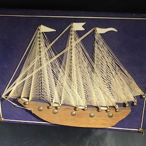 Può includere: Una barca a vela in arte di filo con filo bianco e chiodini dorati su uno sfondo blu. La barca a vela ha tre alberi e uno scafo in legno.