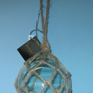 Pode incluir: Um bóia de pesca de vidro azul com uma rede de corda natural, pendurado em um gancho preto. A bóia tem uma etiqueta que diz "Robert's Home Collection".