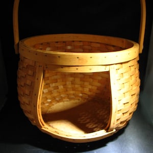 Może przedstawiać: Jasnobrązowy, tkany kosz o zaokrąglonym kształcie i uchwycie. Kosz ma otwór z jednej strony i jest wykonany z cienkich pasków drewna splecionych ze sobą. Kosz jest pusty i stoi na ciemnym tle.