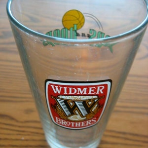 Puede incluir: Un vaso de vidrio transparente con un logotipo rojo, blanco y negro que dice "Widmer Brothers". El logotipo tiene una "W" estilizada con espigas de trigo a cada lado.