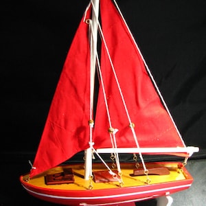 May include: A miniature sailboat model with a red hull and sails. The boat features a white mast and rigging, and is mounted on a white stand. The deck is yellow with brown accents. A decorative nautical item.