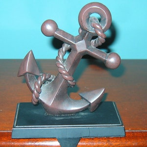 May include: A brown metal anchor stocking holder with a black base and a rope wrapped around the anchor. The stocking holder is sitting on a wooden surface.
