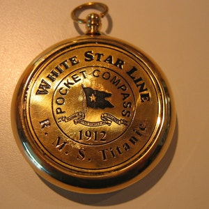 Puede incluir: Br&uacute;jula de bolsillo dorada con una imagen en blanco y negro de una bandera y el texto "White Star Line Pocket Compass R.M.S. Titanic 1912".