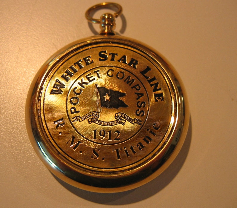 Puede incluir: Br&uacute;jula de bolsillo dorada con una imagen en blanco y negro de una bandera y el texto "White Star Line Pocket Compass R.M.S. Titanic 1912".