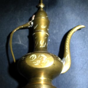 Puede incluir: Una jarra de latón vintage con un pico largo y curvo y un asa. La jarra presenta diseños grabados ornamentados y un cuerpo bulboso. El color general es un oro antiguo cálido.