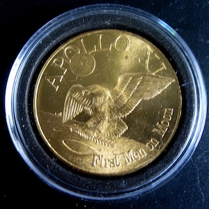 Puede incluir: Una moneda de color dorado con las palabras "Apollo 11" y "First Men on Moon" inscritas alrededor del borde. La moneda presenta una imagen estilizada de un águila con las alas extendidas.